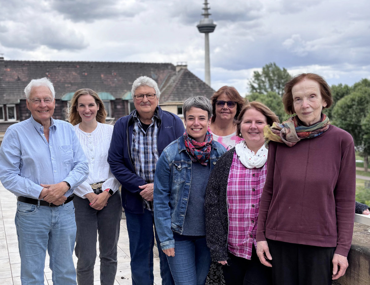 Sieben Personen unterschiedlichen Alters stehen nebeneinander im Freien vor einem Gebäude mit Schindeldach und einem Fernsehturm im Hintergrund