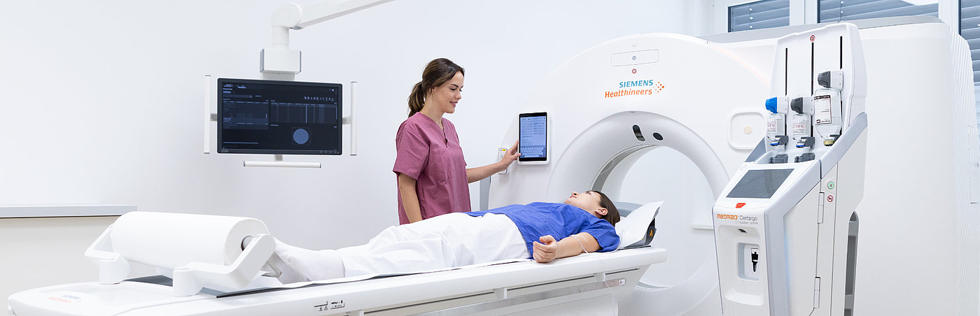 Patientin liegt auf der Liege eines Computertomographen, während eine medizinische Fachkraft die Untersuchung vorbereitet.