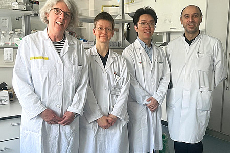 Vier Personen in weißen Laborkitteln stehen in einem Laborraum mit Schränken und Laborgeräten