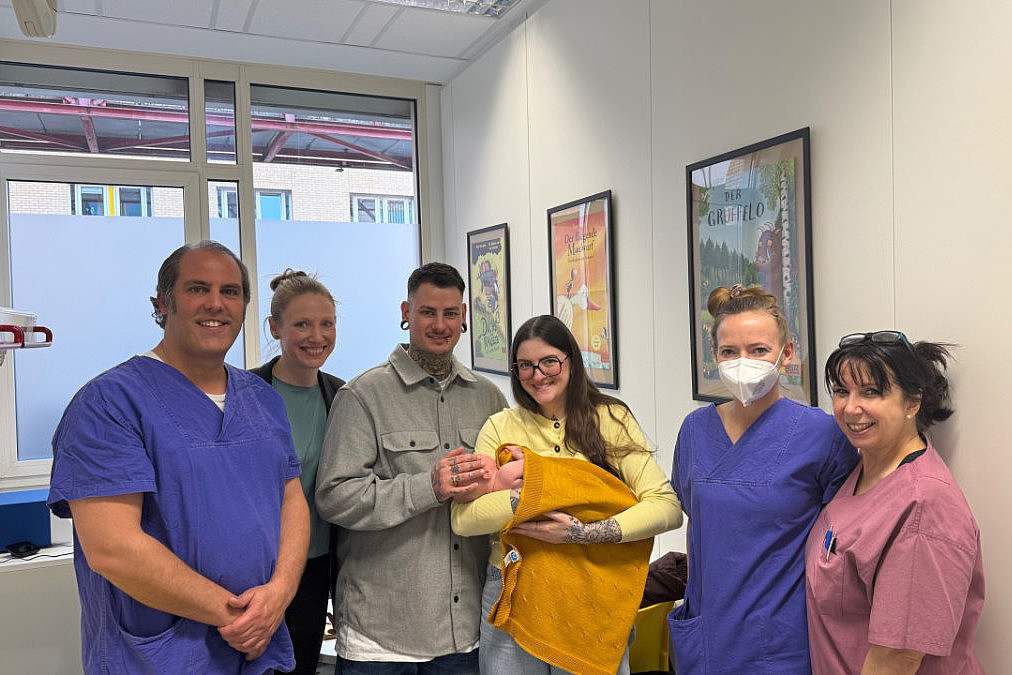 Sechs Personen in einer hellen Praxis: drei in blauer medizinischer Kleidung, drei Frauen. Eine der Frauen hält im Arm ein Säugling; alle lächeln in die Kamera, im Hintergrund Fenster und Bilder an der Wand.
