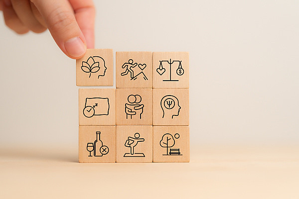 Auf dem Bild sind neun Holzklötzchen gezeigt, auf die jeweils ein Aspekt von Gesundheitsförderung aufgezeichnet ist, sowie eine Hand, die ein Klötzchen hält.