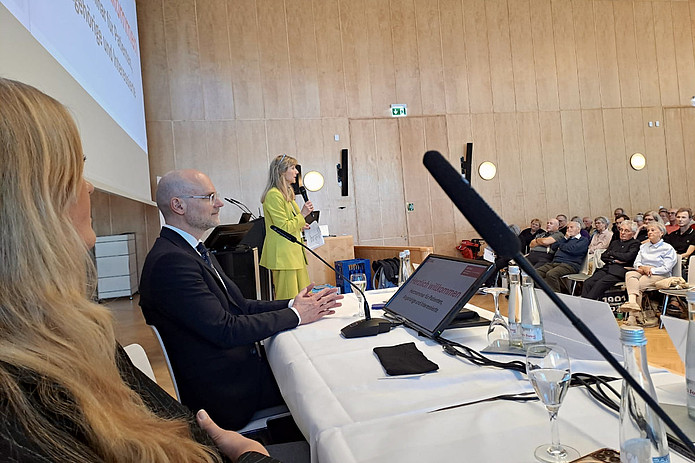 Frau in gelbem Anzug hält Vortrag vor Publikum in hellem Konferenzraum mit Holzvertäfelung