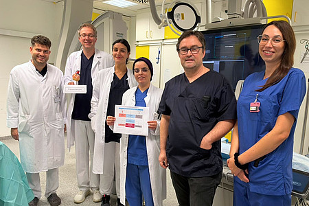 Team der Kardiologie der UMM in einem Herzkatheterlabor. Eine Mitarbeiterin hält eine Übersicht zur Studie, ein Kollege ein Modell mit der Aufschrift „Frauenherzen“.