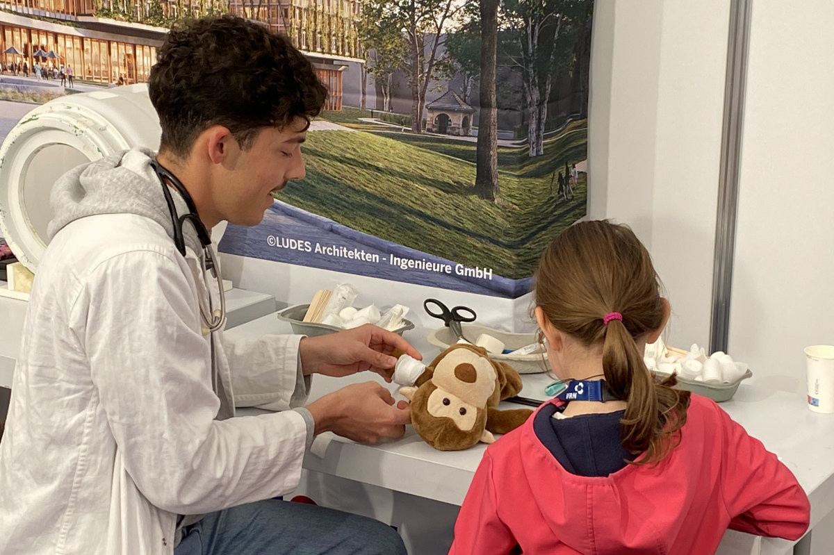 Ein Mann im weißen Kittel mit Stethoskop behandelt sitzend ein Stofftier auf einem Tisch vor einem Mädchen mit rotem Kapuzenpullover