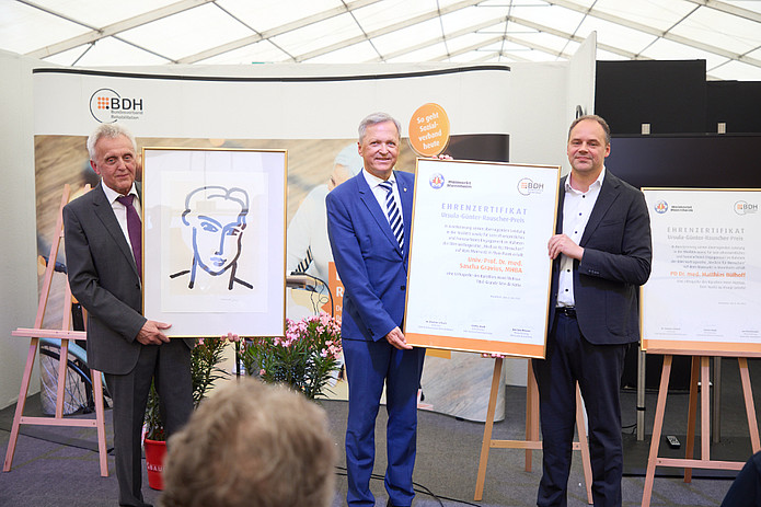 Drei Männer im Anzug auf Bühne halten Urkunde in den Händen.