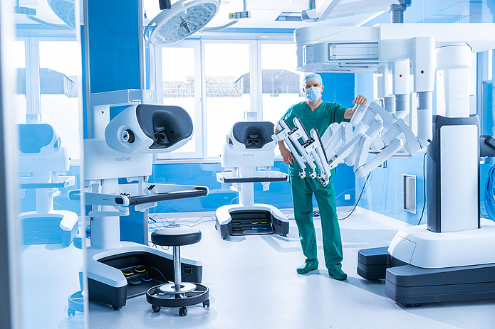 Chirurg im OP-Saal neben einem robotergestützten Operationssystem und weiteren medizinischen Geräten.