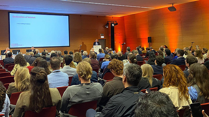 Publikum in einem Konferenzsaal hört einem Redner an einem Podium mit großem Bildschirm zu