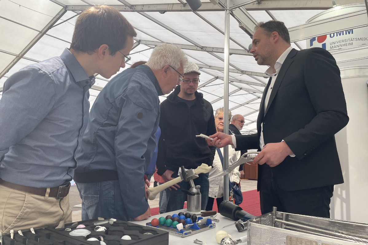 Mehrere Personen stehen an einem Tisch mit medizinischen Modellen und Instrumenten unter einem Zelt, eine Person im Anzug hält ein Modell eines Knochens mit einer Zange