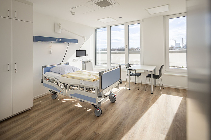 Modern ausgestattetes Patientenzimmer mit Pflegebett, medizinischer Versorgungseinheit und Sitzbereich am Fenster.