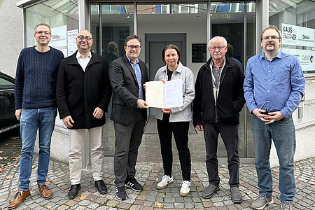 personengruppe vor hauseingang, eine für die förderung symbolhafte urkunde wird übergeben