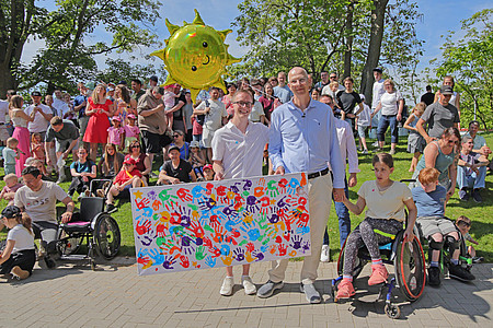 Große Gruppe von Erwachsenen und Kindern steht im Freien zusammen und präsentiert ein bunt gestaltetes Gemeinschaftskunstwerk mit Handabdrücken.