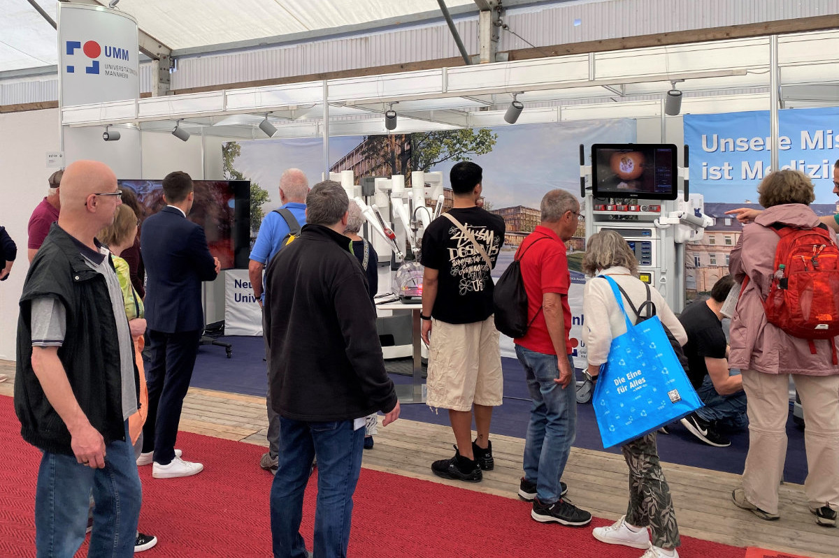 Menschen stehen vor einem Messestand mit medizinischer Ausrüstung und Monitoren unter einem weißen Zeltdach