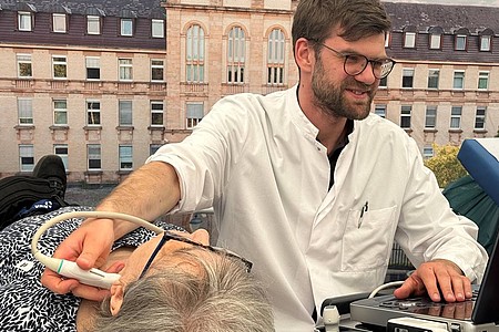 Mann in weißem Kittel führt Ultraschalluntersuchung am Hals einer liegenden älteren Frau durch, im Hintergrund ein historisches Gebäude