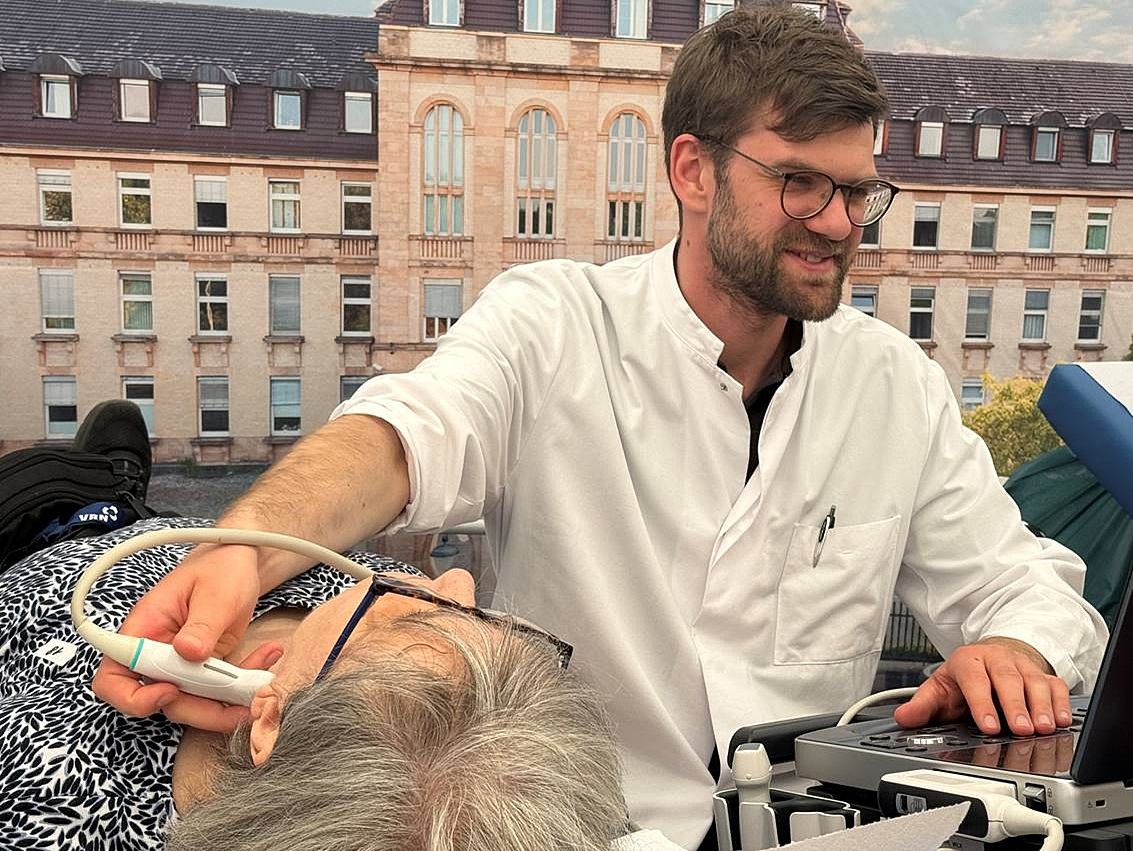 Mann in weißem Kittel führt Ultraschalluntersuchung am Hals einer liegenden älteren Frau durch, im Hintergrund ein historisches Gebäude