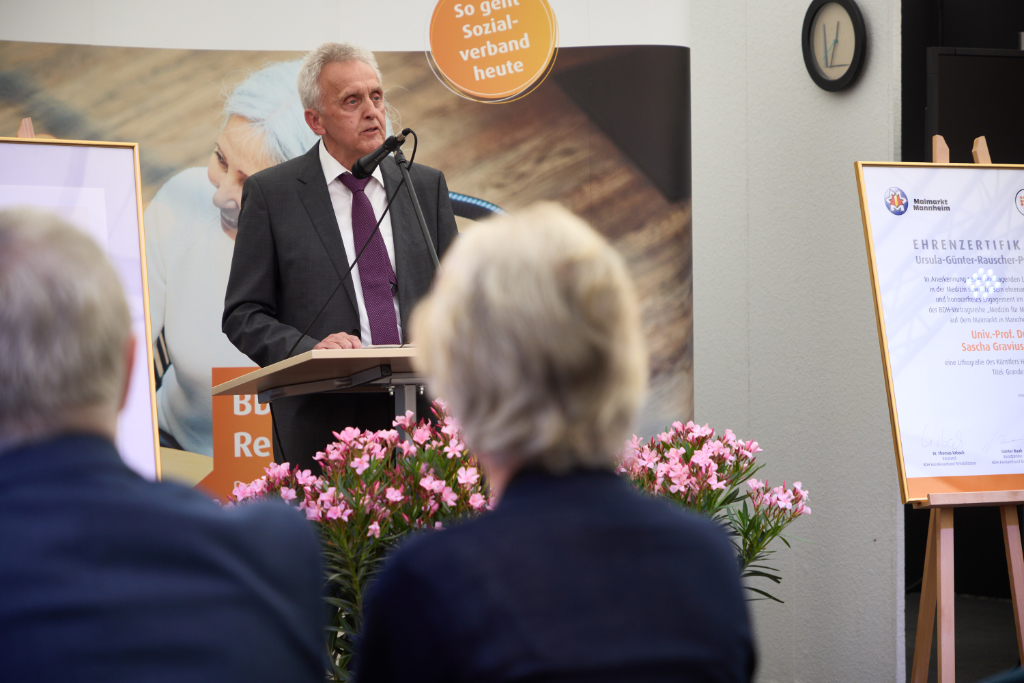 Mann im schwarzen Anzug spricht auf Bühne vor Publikum