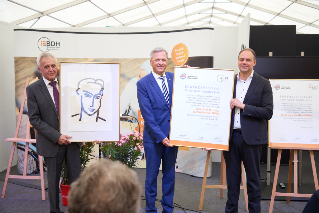 Drei Männer im Anzug auf Bühne halten Urkunde in den Händen.