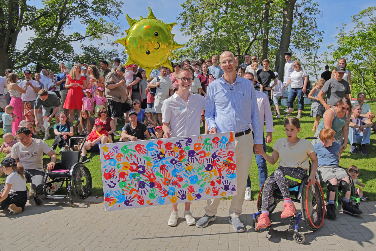 Große Gruppe von Erwachsenen und Kindern steht im Freien zusammen und präsentiert ein bunt gestaltetes Gemeinschaftskunstwerk mit Handabdrücken.