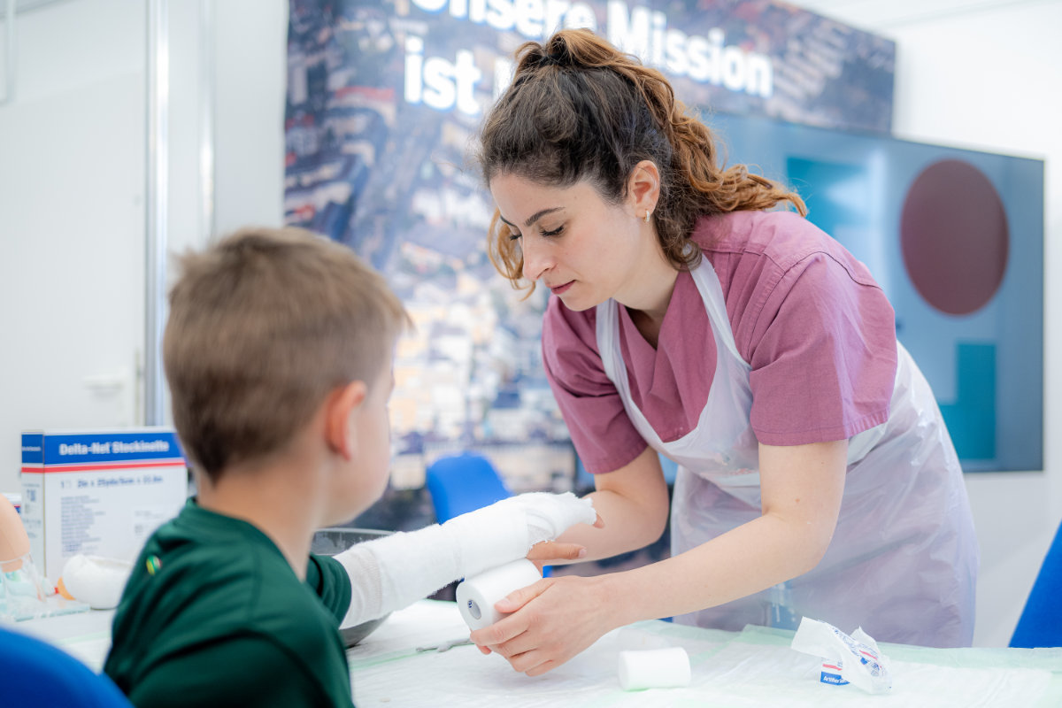 Frau in rosa Oberteil und weißer Schürze wickelt Verband um den ausgestreckten Arm eines Jungen mit grünem Shirt